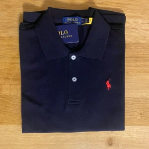 Marinblå pikétröja från Polo Ralph Lauren - Klassisk marinblå pikétröja från Polo Ralph Lauren med röd broderad logga på bröstet. Tröjan har krage, knappar framtill och är tillverkad i bomull. Perfekt för en stilren och tidlös look.