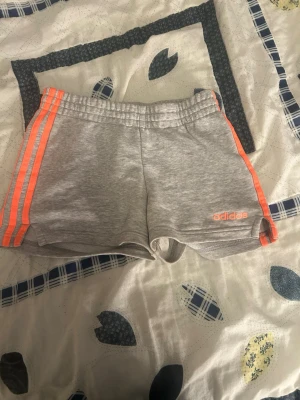 Grå sportshorts från Adidas - Snygga grå shorts från Adidas med orangea ränder längs sidorna och Adidas-logga framtill. Mjukt och stretchigt material med resår i midjan för skön passform. Perfekta för träning eller chill dagar.
