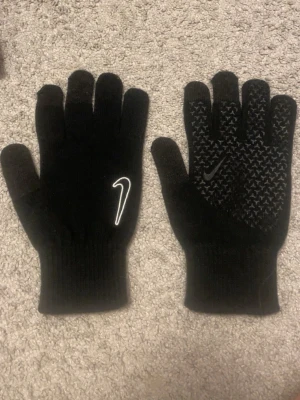 Svarta Nike handskar med grepp - Svarta handskar från Nike med vit logga på ovansidan och greppmönster i gummi på insidan. Perfekta för kyliga dagar och sportiga aktiviteter. Stickad mudd för extra komfort och passform.
