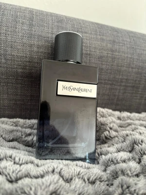 Yves Saint Laurent parfym 100ml - Snygg parfymflaska från Yves Saint Laurent i mörkgrått glas med metallisk etikett och svart lock. Flaskan har en stilren, rektangulär form och YSL-loggan på locket. Perfekt för dig som gillar exklusiva dofter och elegant design.