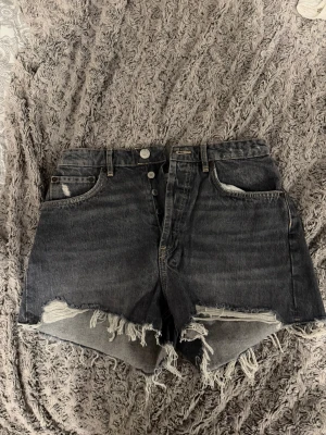 Svarta högmidjade jeansshorts Zara - Säljer ett par svarta högmidjade jeansshorts med råa, fransiga kanter och klassisk femficksdesign. Shortsen har knappar i gylfen och en loose passform. Perfekta för sommaren och ger en avslappnad vibe.