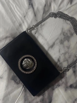 Svart axelväska från Versus Versace - Snygg svart axelväska i läder från Versus Versace med silverfärgad kedjerem. Väskan har ett stort lejonhuvud i silver och strass framtill samt diskret logga på baksidan. Perfekt för dig som gillar statement-detaljer och exklusiv känsla. 