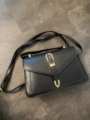 Svart axelväska med gulddetaljer - Stilren svart axelväska i slätt konstläder med justerbar axelrem och snygga guldfärgade metalldetaljer. Väskan har ett lock med spänne och magnetlås samt ett praktiskt innerfack. Perfekt för dig som gillar klassisk och tidlös design.