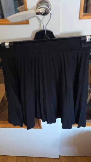 Svart plisserad kortkjol - Snygg svart plisserad kortkjol med bred midjeresår. Kjolen har klassiska veck och är perfekt för en trendig och stilren look. Passar till många olika outfits och är enkel att matcha.