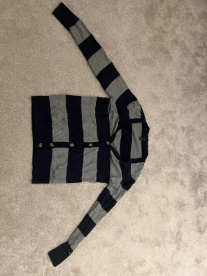 Randig kofta från Tommy Hilfiger - Snygg kofta från Tommy Hilfiger i marinblått och grått med breda horisontella ränder. Långärmad modell med knäppning framtill och rund halsringning. Perfekt för lager-på-lager och enkel att matcha med jeans eller kjol.