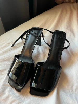 Svarta lackpumps med rem från H&M - Säljer ett par svarta pumps i lack från H&M med fyrkantig tå och bred rem över foten. Skorna har en smal ankelrem och klack, vilket ger en trendig och stilren look. Perfekta för dig som gillar minimalistisk design och glansigt material.