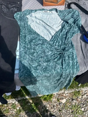 Grön omlottklänning från indiska - Grön omlottklänning från indiska med vitt paisleymönster. Klänningen har v-ringning och korta ärmar, samt en smickrande omlottdesign som knyts i sidan. Perfekt för varma dagar och ger en avslappnad men ändå snygg look.