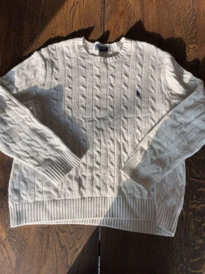 Vit kabelstickad tröja Polo Ralph Lauren - Kabelstickad vit tröja från Polo Ralph Lauren med rund halsringning och klassisk marinblå logga broderad på bröstet. Tröjan har långa ärmar och ribbade muddar vid ärmslut och nederkant. Perfekt för en clean och stilren look.