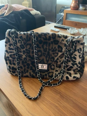 Leopardmönstrad fluffig handväska - Cool handväska med leopardmönster i beige och svart. Väskan har en mjuk, fluffig yta och en lång kedjerem i silver och svart. Stängs med ett vridlås i metall framtill. Perfekt för dig som vill sticka ut med din stil.