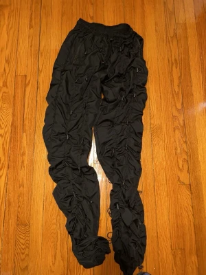 Bunge cord pants cargobyxor MNML ÄKTA - NYSKICK Säljer ett par svarta cargobyxor med unik ruched design och dragsko-detaljer längs hela benen. Byxorna har elastisk midja och flera fickor. Perfekta för dig som vill sticka ut med en streetwear-look.