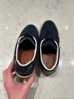 Mörkblå sneakers från Loro Piana - Stilrena mörkblå sneakers från Loror Piana med ovandel i mocka och läder, vita detaljer runt öppningen och klassisk gummisula. Snygg låg profil och diskret logga på innersulan. Perfekta för en clean och modern look.