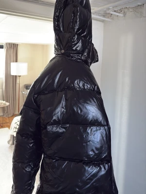 Svart glansig pufferjacka med huva - Säljer en svart pufferjacka med glansig finish och stor huva. Jackan har breda kanaler och en oversized look som ger extra värme. Perfekt för kalla dagar när du vill hålla stilen och samtidigt vara varm.