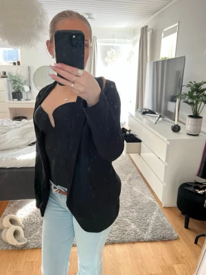 Svart kavaj med klassisk passform - Säljer en svart kavaj med klassisk passform och enkel design. Kavajen har smala slag och är utan synliga knappar framtill. Perfekt att styla över en topp eller skjorta. Materialet ser ut att vara ett mjukt tyg, troligen polyester eller bomullsblandning.