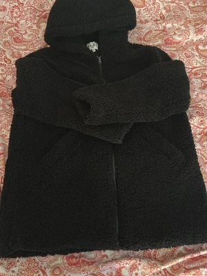 Svart teddyjacka med huva från NA-KD - Mysig svart teddyjacka från NA-KD i storlek 36. Jackan har dragkedja framtill, två sidofickor och en stor huva. Tillverkad i fluffigt teddy-material som håller dig varm och ger en trendig look.