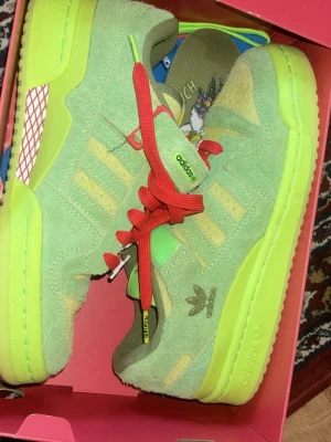 Adidas x Grinch gröna sneakers - Unika Adidas x Grinch sneakers i ljusgrönt mocka med neongula detaljer och röda skosnören. Skorna har klassiska Adidas-ränder och coola Grinch-detaljer på innersulan. Kommer med extra neongula skosnören. Perfekt för dig som vill sticka ut!