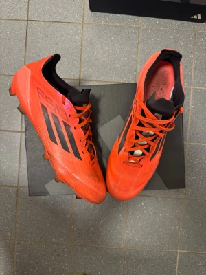 Adidas F50 ”Vivid Horizion” FG - Snygga röda fotbollsskor från Adidas med svarta detaljer och klassiska tre ränder på sidan. Skorna har snörning, låg profil och dobbar för spel på gräs. Ovandelen är i syntetmaterial och insidan är svart. Perfekta för dig som vill ha stil och grepp på planen. KVITTO FINNS FÖR SKORNA, ORIGINAL BOX TILLKOMMER 🤝