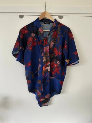 Blommig blus / body med knytband från Zara - Superfin blå mönstrad blus / body / topp från ZARA i storlek S med knytning i halsen och vida korta ärmar. Färgglatt mönster av detaljerade blommor, fjärilar och fåglar i ett lent halvglansigt satin-material. Praktiska tryckknappar i grenen, vilket ger en slät passform. Tyget runt ena tryckknappen har lossnat lite, se bild, men inget som förändrar knäppningen eller syns när det är knäppt. I övrigt i mycket fint skick.  Skickas mot porto, kan även hämtas i Stockholm. 