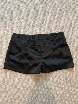 Svarta kostymshorts - Snygga svarta shorts med låg midja och dekorativa knappar fram samt på benen. Shortsen har breda hällor för skärp och två fickor framtill. Ska nämnas att de har ett litet hål på sidan av ena fickan, men inget som man tänker på. Säljer då de är för stora för mig. 