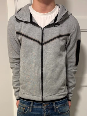 Grå Nike teech fleece - Säljer en grå teech fleece från Nike med svarta detaljer och logga på bröstet. Tröjan har huva, hel dragkedja och fickor på sidorna samt en svart patch på ena ärmen. Perfekt för en avslappnad look. Hör av dig vid minsta intresse! ⭐️💫