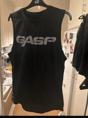 Svart linnetopp från GASP - Säljer en svart ärmlös linnetopp från GASP med tryckt logga framtill och en gul detalj på ryggen. Perfekt för dig som gillar sportig och avslappnad stil. Materialet känns mjukt och lätt, vilket gör den skön att bära.