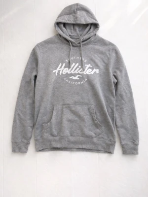 Grå hoodie från Hollister L 14-16 - Snygg grå hoodie från Hollister med vit logga och texten 'Southern California' på bröstet. Klassisk känguruficka framtill, dragsko i huvan och mjukt material som känns skönt mot huden. Perfekt för en avslappnad och trendig stil.