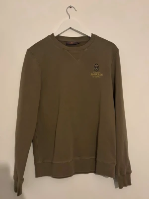 Olivgrön sweatshirt från Morris - Snygg olivgrön sweatshirt från Morris med rund halsringning och broderad logga på bröstet i gult och svart. Tröjan har ribbade muddar vid ärmslut och nederkant samt en klassisk relaxed fit. Perfekt för en avslappnad och stilren look.