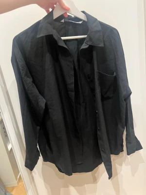 Svart oversized skjorta i bomull - Stilren svart skjorta med klassisk krage och bröstficka. Skjortan har en oversized passform och är tillverkad i mjuk bomull. Perfekt för en avslappnad look och enkel att styla med olika outfits.