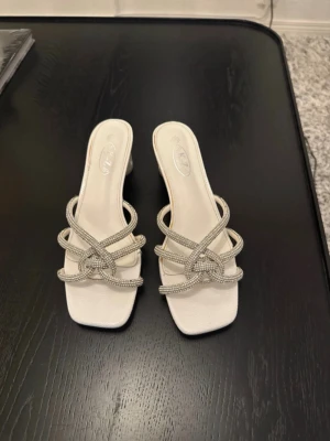 Glittriga klackskor  - Säljer dessa fantastiska vita klackskor (mules) som verkligen sticker ut! De har korsade remmar prydda med glittriga stenar och en riktigt cool, genomskinlig klack med dekorativa detaljer. • Storlek: 40 • Färg: Vit med silverglitter/strass • Klack: Stabil blockklack i genomskinlig design • Skick: Sparsamt använda (se bild). • Märke: K-Z Dessa är perfekta för bröllop, sommarfesten eller när du bara vill liva upp en outfit! 📦 fraktar snabbt