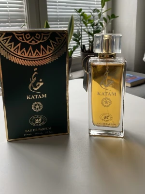 Katam Eau de Parfum 100ml - Exklusiv parfym från Katam i elegant rektangulär glasflaska med genomskinlig kork och guldfärgade detaljer. Doften kommer i en lyxig mörkgrön och guld kartong. Flaskan rymmer 100 ml Eau de Parfum och har en stilren, modern design.