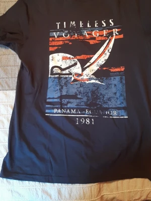 Svart t-shirt Regatta med tryck - Cool svart t-shirt från Regatta med stort grafiskt tryck i vitt, blått och rött framtill. Motivet har texten 'Timeless Voyager', 'Panama Ecuador 1981' och en segelbåt. Perfekt för dig som gillar unik design och marint tema. UNISEX. Storlek för man: S, kvinna: M. Mått på bilderna. 