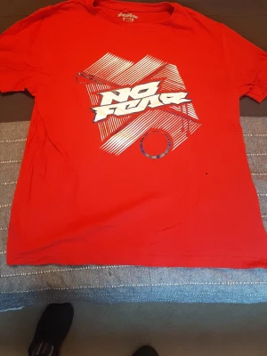 Röd No Fear t-shirt med tryck - Säljer en röd t-shirt från No Fear med stort grafiskt tryck och texten 'No Fear' på bröstet. T-shirten har klassisk passform och korta ärmar. Perfekt för dig som gillar streetwear och vill sticka ut med en färgstark look. Unisex, storlek man: S, kvinna: M. Mått på bilderna. 