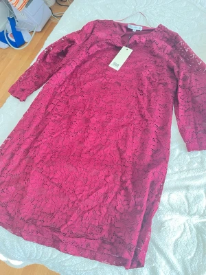 Vinröd spetsklänning från Flash - Vinröd klänning från Flash med blommigt spetsmönster och trekvartsärm. Klänningen har en rund halsringning och är fodrad med ett mjukt tyg. Perfekt för dig som vill ha en elegant och färgstark look.