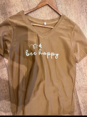 Brun t-shirt med v-ringning - Cool brun t-shirt med v-ringning och vit text 'bee happy' på bröstet, tillsammans med en liten bi-illustration. Perfekt för dig som gillar chill vibes och snygga prints. Materialet känns mjukt och bekvämt, och modellen har korta ärmar.