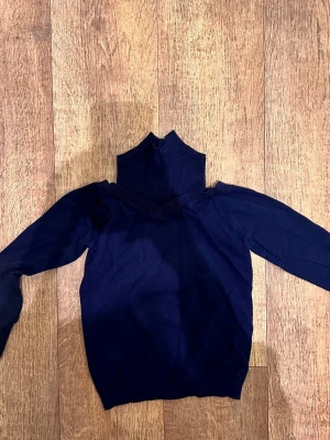 Mörkblå stickad polotröja - Säljer en mörkblå stickad polotröja med hög krage och långa ärmar. Tröjan har ribbstickade detaljer vid kragen och nederkanten, vilket ger en snygg och modern look. Perfekt för dig som gillar stilrena plagg med lite edge.