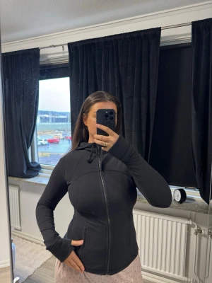 Svart hoodie med dragkedja lululemon - Säljer en svart hoodie med dragkedja och huva. Modellen är figurnära med långa ärmar och fickor på sidorna. Perfekt för dig som gillar en enkel och stilren look. Materialet känns mjukt och bekvämt, och huvan har snören för justering.