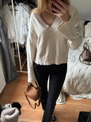 Vit stickad V-ringad tröja - Säljer en krämvit stickad tröja med djup V-ringning och bred ribbad kant vid hals, ärmslut och nederkant. Tröjan har en avslappnad passform och långa ärmar, perfekt för lager på lager. Enkel och stilren design utan synligt märke.