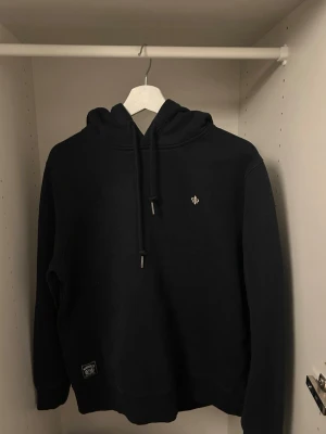 Svart hoodie med broderad detalj - Snygg svart hoodie från Morris med klassisk huva och dragsko. På bröstet finns en liten broderad lilja i vitt. Hoodien har en enkel och stilren design, perfekt för en avslappnad look. Materialet känns mjukt och bekvämt mot huden.