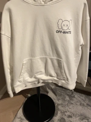 Vit Off-White hoodie med tryck - Vit hoodie från Off-White med svart tryck framtill och stort grafiskt motiv på ryggen. Klassisk känguruficka och huva. Perfekt för dig som gillar streetwear och vill sticka ut med en unik design.