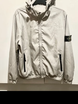 Stone Island Hooded Jacket – grå – Nyskick - Stone Island jacka i mycket fint skick. Endast använd ett fåtal gånger så den är i princip som ny. Stilren beige/grå färg med huva, dragkedja och klassiska Stone Island-badgen på armen. Perfekt vår/höstjacka som passar till allt. Nypris betydligt högre. Skriv vid frågor eller fler bilder.
