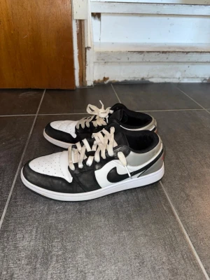 Nike Air Jordan 1 Low svart/vit - Säljer ett par Nike Air Jordan 1 Low sneakers i svart och vitt med klassisk swoosh på sidan. Skorna har snörning, perforerad tåbox och lågt skaft. Insidan är svart med röd detalj på plösen. Perfekta för dig som gillar streetwear och ikonisk design.1,1