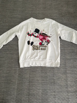 Vit sweatshirt med snögubbe från Lindex  - Vit sweatshirt med tryck av en snögubbe och varm choklad, samt texten 'Stay warm, chill out' framtill. Tröjan har rund hals, långa ärmar och ribbade muddar. Perfekt för vinter och jul med sitt gulliga motiv. Bra skick utom en fläck (4 bilden) Säljer då det blivit för litet