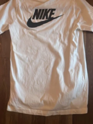 Vit Nike t-shirt med logga - Säljer en vit t-shirt från Nike med stor svart logga på ryggen och mindre tryck med Nike-text och swoosh på bröstet. Klassisk rund hals och korta ärmar. Perfekt för dig som gillar sportig och clean stil.