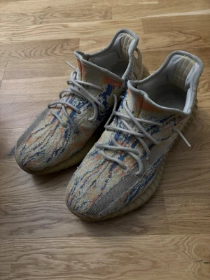 Yeezy Boost 350 V2 sneakers beige/multicolor - Säljer ett par Yeezy Boost 350 V2 sneakers i beige med färgstarka detaljer i blått, orange och gult. Skorna har en stickad ovandel, genomskinlig sula och klassiska Yeezy-formen med rund tå och lågt skaft. Perfekta för dig som gillar unik design och streetwear.