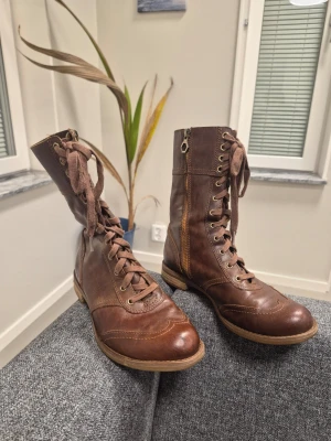 Timberland Savin Hill Mid Kängor Brun EU 37.5 - Snygga bruna stövletter i äkta läder med snörning framtill och dragkedja på sidan. De har en klassisk rund tå och låg klack, samt detaljerade sömmar som ger en vintagekänsla. Perfekta för dig som gillar en tidlös och cool stil.