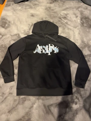 Svart AMIRI hoodie med drakmotiv - Säljer en svart hoodie från AMIRI med stor huva och känguruficka. På bröstet finns ett stort AMIRI-tryck dekorerat med blåa drakar. Hoodien har ribbade muddar och snörning i huvan. Perfekt för dig som gillar streetwear och coola detaljer.