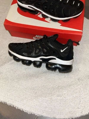Nike Air Vapormax Plus svarta sneakers - Nike Air Vapormax Plus sneakers i svart med vit sula och transparenta luftbubblor. Ovandelen är i textil och syntet med ikoniska detaljer och små vita swoosh-loggor. Skorna har en sportig och futuristisk look med snörning och låg profil.