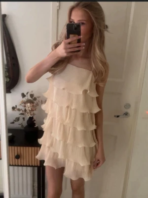 Champagnefärgad volangklänning - Säljer en kort klänning i champagnefärg med flera lager volanger och tunna axelband. Klänningen har en luftig och festlig design som ger ett snyggt och ungdomligt intryck. Perfekt för dig som gillar att sticka ut med stilrena detaljer.