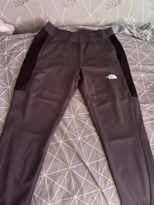 Grå mjukisbyxor från The North Face - Mörkgrå mjukisbyxor från The North Face med svarta paneler längs sidorna. Byxorna har resår i midjan och logga tryckt på både fram- och baksidan. Perfekta för chill eller träning, tillverkade i mjukt och stretchigt material. Köpt 2025 använd få tal gånger 