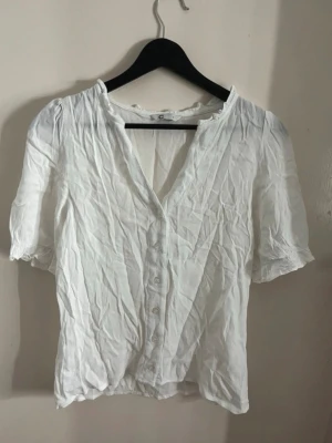 Vit kortärmad blus med knappar - Söt vit blus med korta puffärmar och knäppning framtill. Blusen har en lätt v-ringning och små volangdetaljer vid halsringningen. Tillverkad i ett tunt, luftigt tyg som ger en avslappnad känsla. Perfekt att matcha med jeans eller kjol. Super skön till i sommar. Fin vit blus. Skrynklig nu men vid strykning blir den perfekt igen. Super snygg och söt. Sitter bra på och har en bra passform. Jätte fin. 
