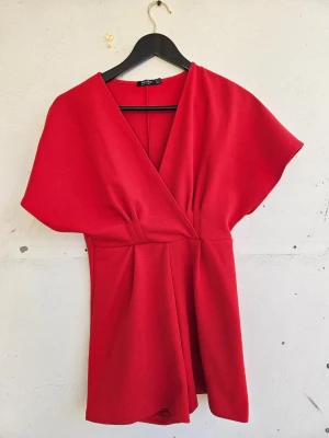 Röd kortärmad jumpsuit med omlott - Snygg röd jumpsuit med korta vida ärmar och omlott-ringning från Bershka i storlek S. Lite sliten i sömmen framtill som man ser på sista bilden, annars fint skick.